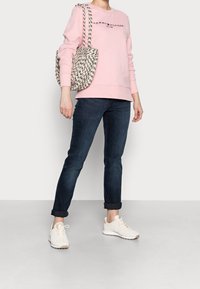 Žena v růžové mikině Tommy Hilfiger, tmavých zastrčených džínách, bílých teniskách a s černobílou vzorovanou taškou přes rameno.