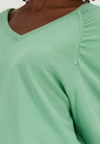 Lichtgroene v-hals trui met geribde halslijn en subtiele pofmouwen. Zachte stof met een gladde textuur en gestikte details op de naden.