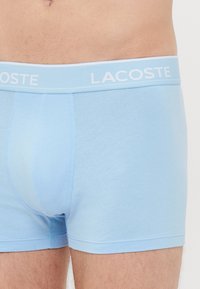 Torso masculino con calzoncillos Lacoste de color azul claro, con el logo visible en la banda elástica, mostrando un primer plano del ajuste de la ropa interior y la textura de la tela.