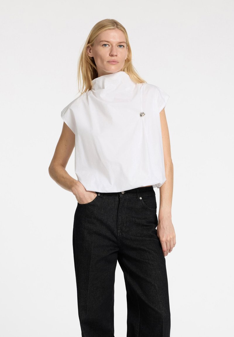 Top in cotone bianco con colletto alto e maniche corte, con un accento argentato sul petto, abbinato a pantaloni in denim neri.