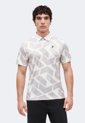 Muškarac obučen u sivu i bijelu polo majicu geometrijskog uzorka s tri dugmeta i crne hlače, stoji nasuprot jednostavnoj pozadini.