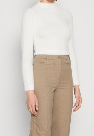 Femme portant un haut blanc ajusté à manches longues et un pantalon beige taille haute, debout devant un fond uni.