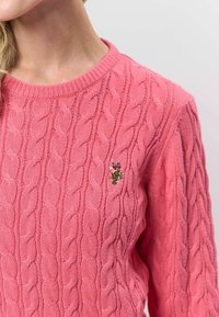 Lyserød kabelstrikket sweater med rund halsudskæring. Har en struktureret vævning og et lille broderet detalje på brystet.