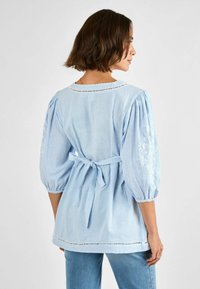 Blusa a righe blu chiaro con maniche a sbuffo, vita legata e ricami decorativi sulle maniche. Ha un collo rotondo e rifiniture in pizzo.