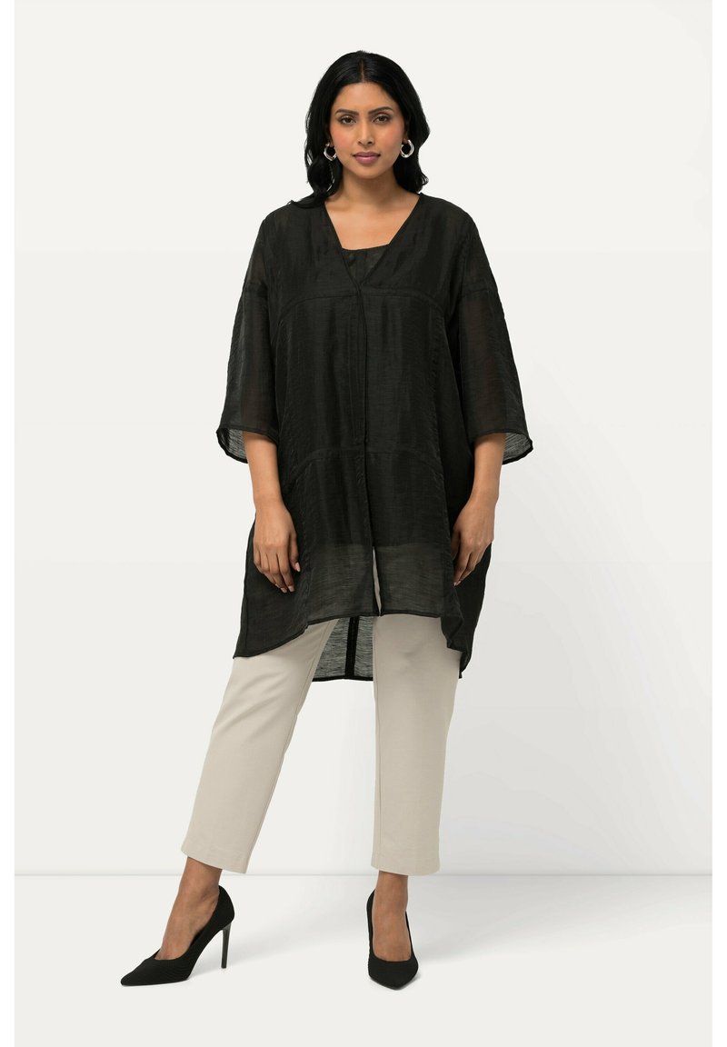 Tunic nera, ampia e svasata con scollo a V e maniche larghe, realizzata in un tessuto testurizzato. Indossata sopra pantaloni beige chiaro e tacchi neri.