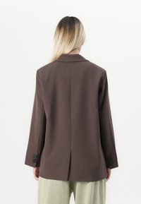 Personne avec de longs cheveux blonds portant un blazer marron foncé et un pantalon clair, vue de dos sur fond blanc uni.