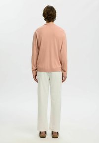 Personne aux cheveux courts et bouclés portant une chemise à manches longues rose clair, un pantalon blanc et des sandales noires, debout de dos sur un fond blanc.