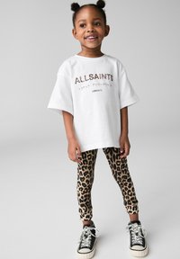 Biała, oversize'owa koszulka z nadrukiem "ALLSAINTS", dopasowane legginsy w panterkę oraz czarne sneakersy z białymi akcentami.