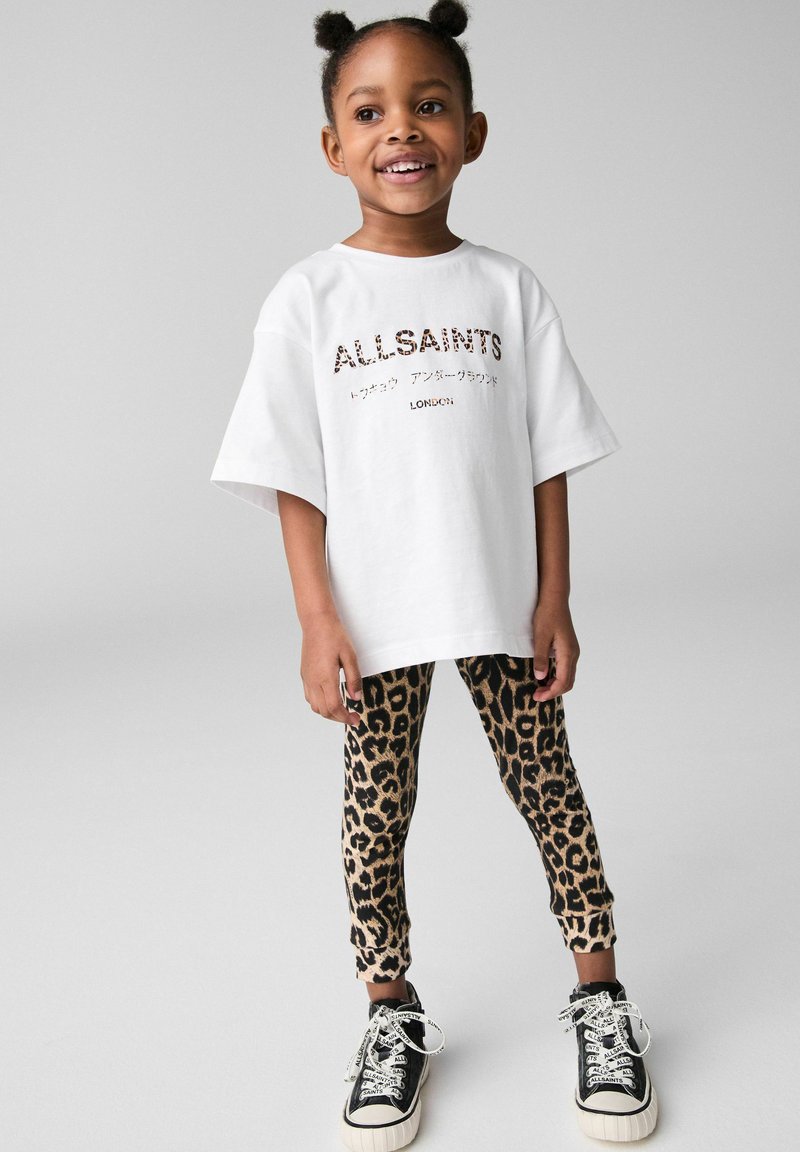 Biała, oversize'owa koszulka z nadrukiem "ALLSAINTS", dopasowane legginsy w panterkę oraz czarne sneakersy z białymi akcentami.