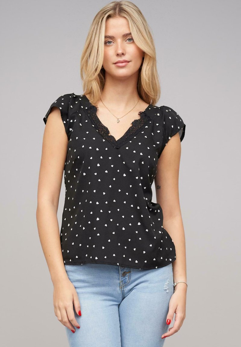 Zwarte blouse met witte hartpatronen, kant V-halslijn, korte pofmouwen en een ontspannen pasvorm, gecombineerd met lichtblauwe spijkerbroek.
