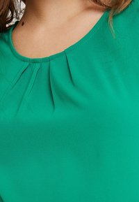 Green blouse with a round neckline and pleated detailing. Made of smooth fabric, featuring light texture and soft draping around the shoulders.