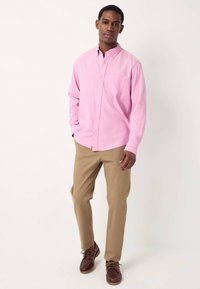 Vaaleanpunainen nappikauluspaita, jossa on rintatasku, yhdistetty beigenvärisiin housuihin ja ruskeisiin nauhakenkiin. Yksinkertainen muotoilu ja pehmeä tekstiili.