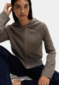 Šedá mikina na zip vyrobená z měkkého materiálu s žebrovanými manžetami a spodním lemem, s kapucí a stříbrným zipem. Nosí se s tmavými džínami.