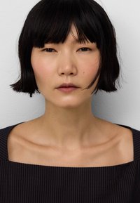 Portret osoby z krótkimi czarnymi włosami, gładką skórą, ubraną w ciemną, fakturowaną bluzkę z kwadratowym dekoltem i subtelnymi pionowymi paskami.