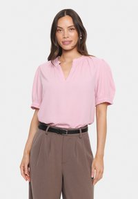 Rosa Bluse mit kurzen Puffärmeln, gerafftem Ausschnitt und strukturiertem Stoff, kombiniert mit hochgeschnittenen, taillierten braunen Hosen und einem schwarzen Gürtel.