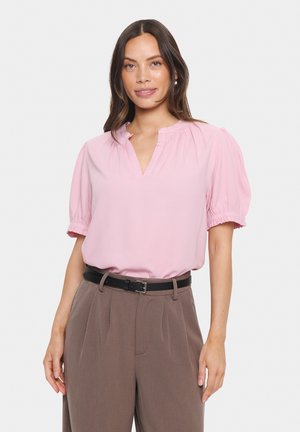 Rosa Bluse mit kurzen Puffärmeln, gerafftem Ausschnitt und strukturiertem Stoff, kombiniert mit hochgeschnittenen, taillierten braunen Hosen und einem schwarzen Gürtel.