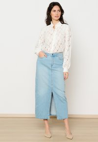 Bela čipkasta bluzica z visokim ovratnikom in sprednjim gumbom, v kombinaciji s svetlo modrim jeans krilom z razporkom spredaj. Čevlji nevtralne barve.