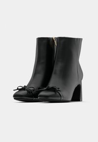 Botas de tornozelo em couro preto com bico arredondado, apresentando um fecho lateral e laços de fita preta na frente. Salto quadrado para suporte.