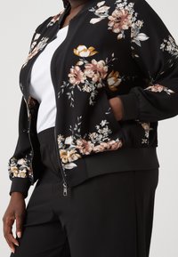 Černá bomber bunda s květinovým vzorem v růžové, oranžové a bílé; má zipové zapínání a žebrované manžety; nošena přes bílé tričko.