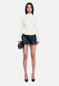 Weißes Spitzenoberteil mit langen Ärmeln und strukturiertem Muster, kombiniert mit einem schwarzen Leder-Minirock. Das Model hält eine schwarze Clutch und trägt schwarze High Heels.