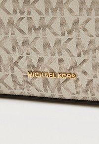Beige gestructureerde stof met een herhalend logo patroon en gouden "MICHAEL KORS" branding. Heeft een gladde afwerking en subtiele details.