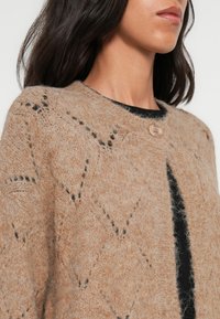 Cardigan beige en tricot avec un col rond, présentant un motif en losange et une fermeture à un bouton marron. Tissu doux et texturé.