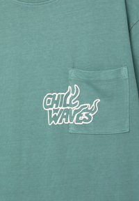Turkis bomulds t-shirt med rund hals, med et hvidtrykt logo "CHILL WAVES" og en lille brystlomme.