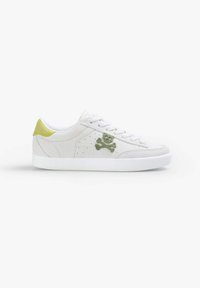 Białe zamszowe sneakersy z zielonym emblematem czaszki i skrzyżowanych kości, perforowanymi akcentami oraz teksturowaną białą gumową podeszwą.