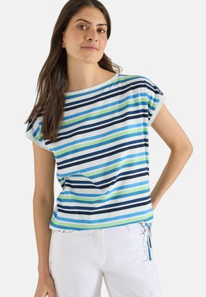 Femme aux longs cheveux bruns portant un t-shirt à manches courtes à rayures horizontales multicolores et un pantalon blanc, debout devant un fond uni.