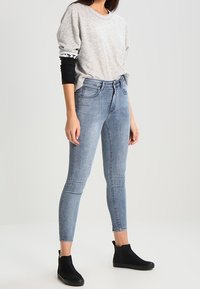 Mujer vistiendo un suéter ligero de mangas abullonadas gris claro, capas de camisetas de manga larga en blanco y negro, jeans ajustados azul claro y botines negros, de pie.