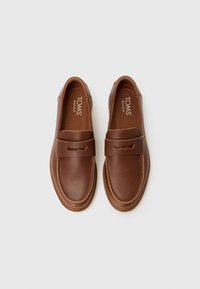 Mocasines de cuero marrón con suela plana, que presentan un diseño cosido y una correa ancha en la parte delantera. Superior texturizado con plantilla de marca.