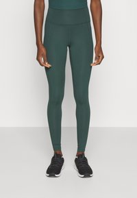 Leggings taille haute, vert foncé, fabriqués à partir d'un tissu extensible avec un ajustement serré. Présentent des coutures plates et sont associés à des chaussures de sport noires.