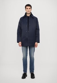 Navy blue waterproof jacket with a straight cut, snap buttons, side pockets, and a collar. Worn over a dark sweater and blue jeans.