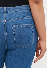 Nærbilde av en person som har på seg blå denimjeans med en baklomme og en svart topp, hvor høyre hånd henger avslappet ved siden av.