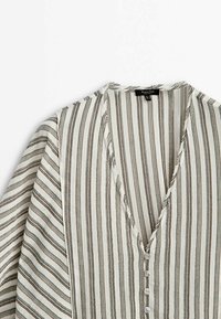 Blouse blanche à fines rayures verticales noires, col en V, quatre boutons sur le devant et manches longues à coupe ample.