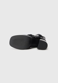 Zwarte hoge hak schoen met een gladleren bovenzijde en een gestructureerde rubberen zool. Heeft een brede band met zilverkleurige hardware-accenten.