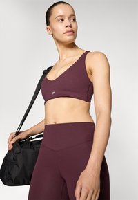 Μπορντό αθλητικό σουτιέν με φαρδύς ώμους και βαθύ V ντεκολτέ, σε συνδυασμό με ψηλόμεσα κολάν στο ίδιο χρώμα; το μοντέλο κρατά μια μαύρη τσάντα γυμναστικής.