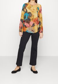 Blouse à manches longues multicolore avec un motif floral dans des tons de jaune, bleu, rose et marron, associée à un pantalon noir à jambes droites.
