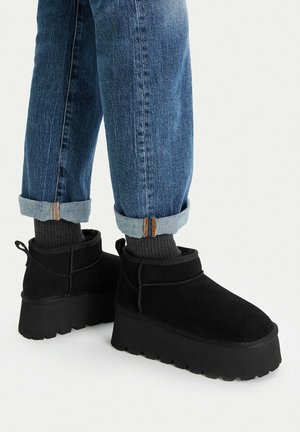 Legs wearing cuffed blue jeans, dark gray socks, and black platform ankle boots with thick ridged soles on a white background.