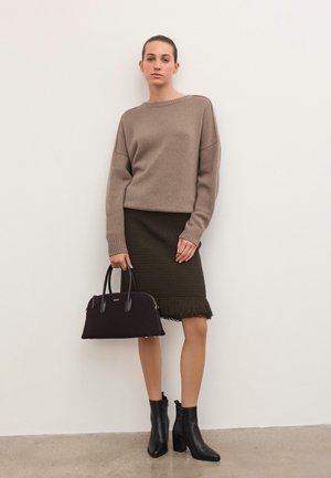Woman wearing a beige sweater and dark green knitted skirt with fringe, holding a black handbag and wearing black ankle boots against white wall.