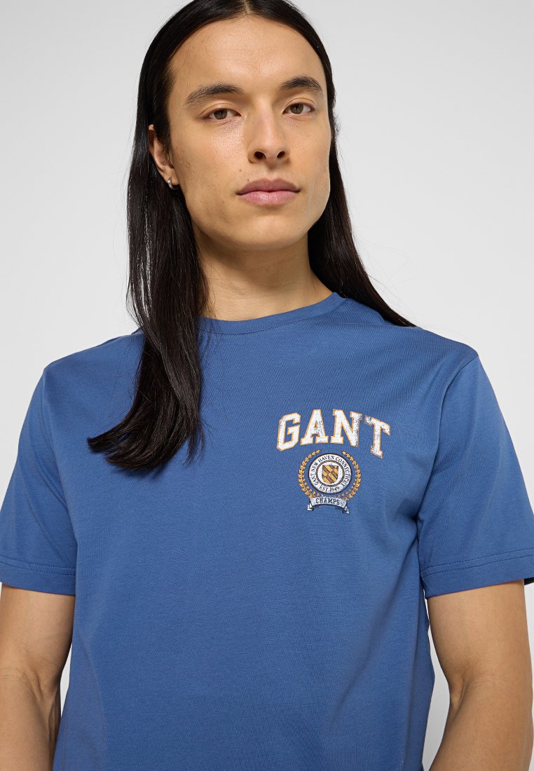 Blaues Baumwoll-T-Shirt mit Rundhalsausschnitt und gesticktem Logo. Ausgestattet mit einem runden Emblem und Text in weißen und goldenen Stichen. Kurzarm.