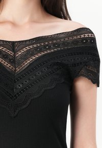 Melna off-the-shoulder blūze ar mežģīņu apkakli, kurā ir sarežģīti raksti un ribota tekstūra, apdarināta ar viļņotām malām.