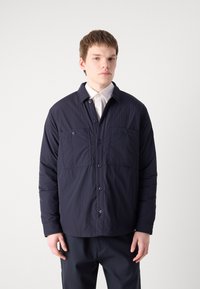 Chaqueta acolchada azul marino con un diseño de cuello, dos bolsillos frontales y cierre de botón. Fabricada con un tejido ligero y texturizado.