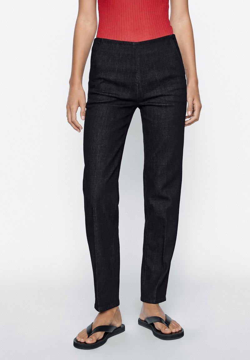 Pantalon en denim noir à coupe slim, taille haute et devant plat, porté avec des tongs noires et un haut rouge en maille côtelée.