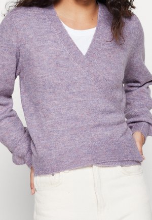 Personne portant un pull doux à col en V lavande par-dessus un haut blanc, associé à un pantalon taille haute de couleur blanc cassé, les mains dans les poches.