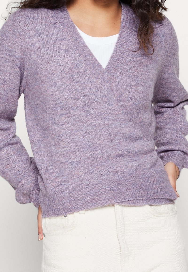 Personne portant un pull doux à col en V lavande par-dessus un haut blanc, associé à un pantalon taille haute de couleur blanc cassé, les mains dans les poches.