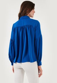 Blusa di satin blu con retro plissettato, colletto e polsini con bottoni, caratterizzata da maniche a sbuffo lunghe e una vestibilit1 comoda. Indossata con pantaloni chiari.