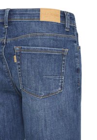 Vue arrière d'un jean en denim bleu montrant la poche arrière droite, les passants de ceinture et une étiquette en cuir beige de la marque "PULZ JEANS" à la taille.