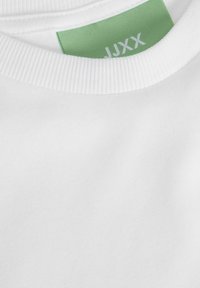 Bílý bavlněný T-shirt s kulatým výstřihem a žebrovaným lemem. Obsahuje zelenou látkovou etiketu s bílým textem uvnitř oblasti výstřihu.