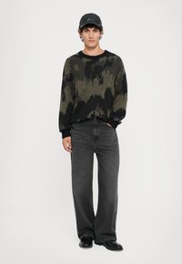 Groene en zwarte geblokte trui, relaxte pasvorm, gecombineerd met donkergrijze brede jeans en een zwarte pet. Model staat tegen een witte achtergrond.
